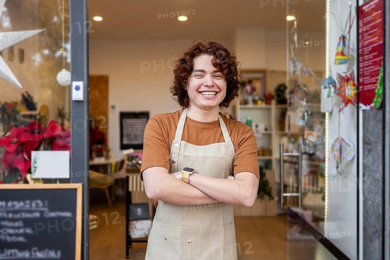 A cheerful young shop assistant in an apron stands with crossed arms at the entrance of an organic products store, inviting customers into the cozy, eco-friendly boutique
