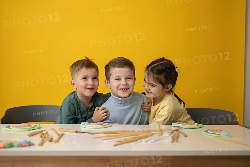 Three smiling children engage in a creative play lesson, surrounded by colorful clay and tools on a bright yellow backdrop, emphasizing joyful learning and creativity