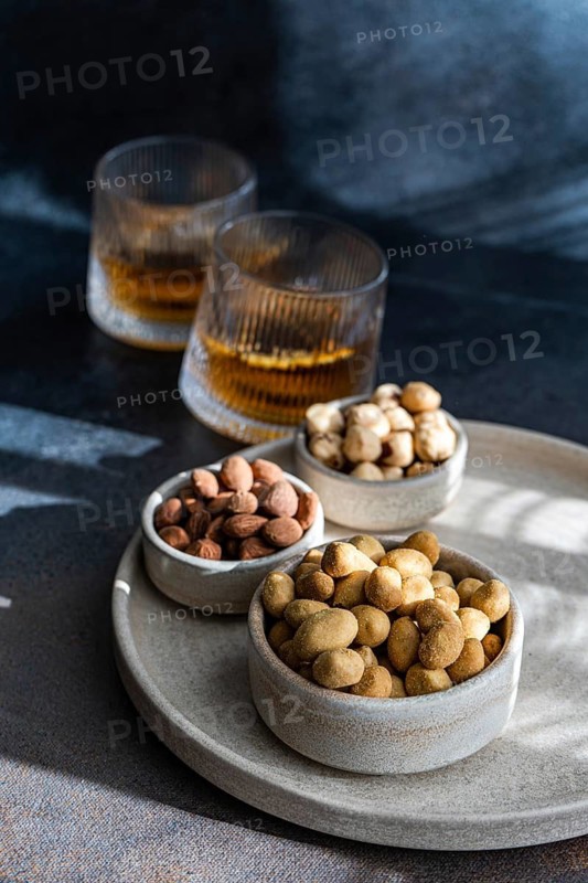 A selection of spiced cognac cocktails, with cinnamon and star anise, accompanied by various nuts including almonds, peanuts, and hazelnuts, elegantly presented on a textured, gray ceramic surface