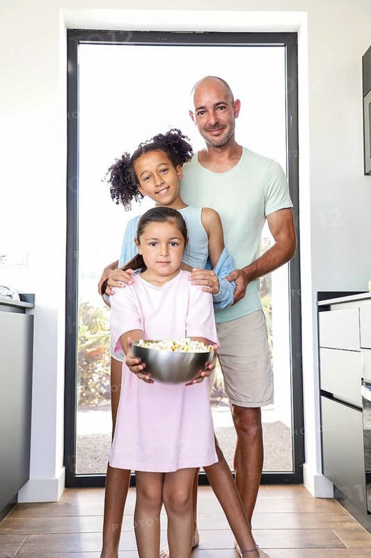 A joyful father with his two daughters enjoying a day at home. The younger child holds a bowl of popcorn, creating a warm and inviting family atmosphere