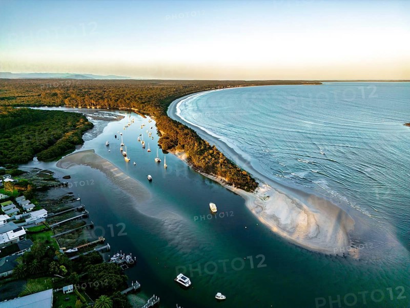 A stunning aerial view captures a serene coastal landscape with a curving coastline, calm river, and anchored boats amid lush greenery, offering a peaceful coastal scene in Jervis Bay
