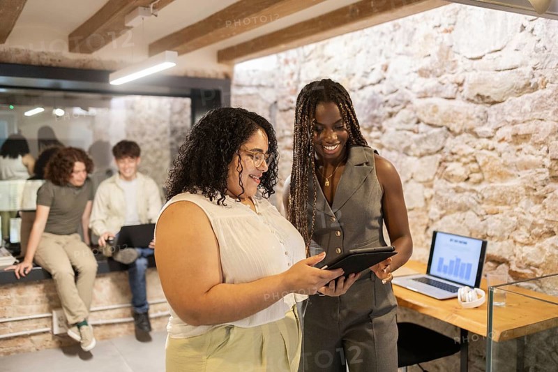 A diverse group of coworkers collaborate in a modern office. Two women stand discussing ideas with a tablet, while two men in the background work on a laptop