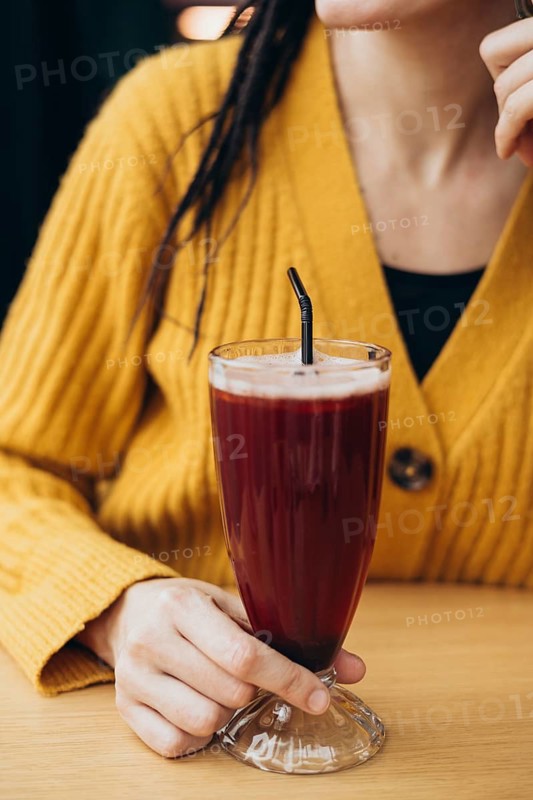 A woman in a yellow sweater holds a tall glass with a refreshing coffee and a straw She sits at a wooden table, creating a cozy, relaxed atmosphere