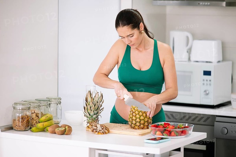 A pregnant woman in a green yoga outfit stands at a kitchen counter slicing a pineapple. The scene highlights wellness, nutrition, and healthy habits during pregnancy