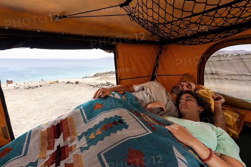 A blind man rests in a motorhome with his partner, wrapped in a colorful blanket, gazing out at the serene ocean. The interior is warm and welcoming, with a view of the beach