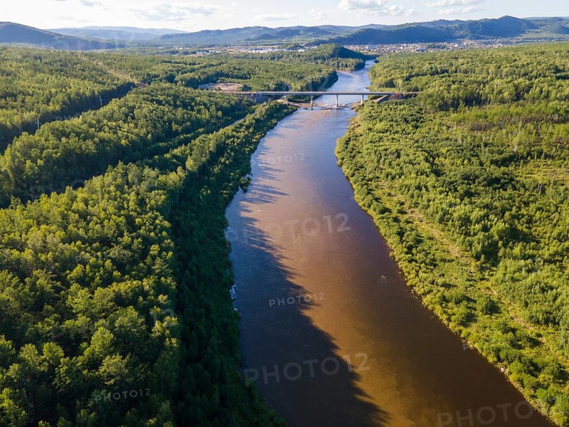 A serene view of a winding river surrounded by lush greenery in Vladivostok, Russia. The scene captures the tranquil beauty of nature with rolling hills in the background and a bridge crossing over the water. It showcases the vibrant ecosystem