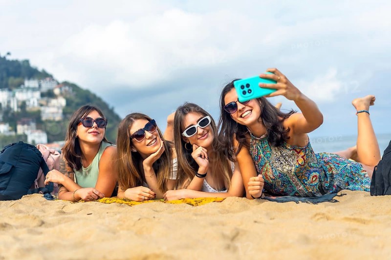 Young women friends lying on warm sand at the beach, smiling and taking a sunny group selfie with a phone, sunglasses and laughter on a summer vacation together