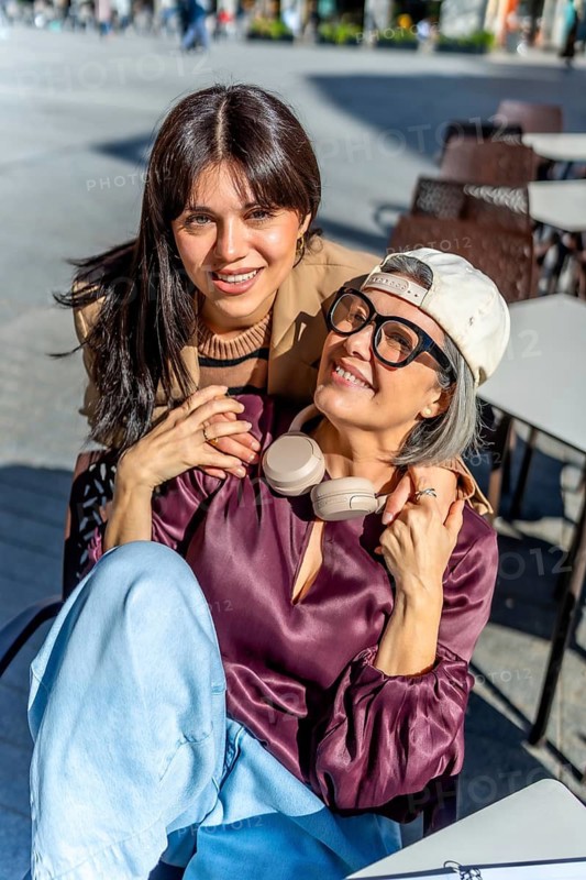 Two womenone younger, one olderembrace and smile at the camera while sitting at an outdoor cafe, sharing a warm, multi generational moment of connection and joy on a sunny day
