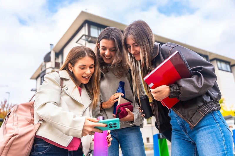 Young women college students checking social media content on a smartphone, sharing a moment and enjoying their friendship outdoors in an urban setting surrounded by campus buildings