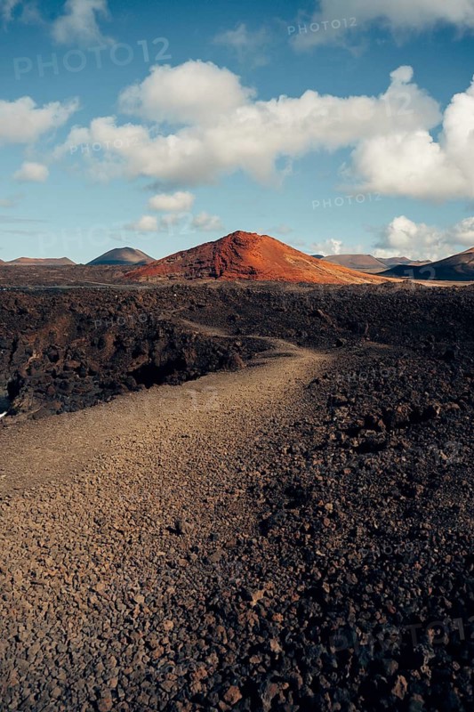 Captivating Lanzarote landscape showcasing volcanic terrain with rusty red hills, rugged lava fields, and a vibrant blue sky dotted with white clouds. A stunning natural scene