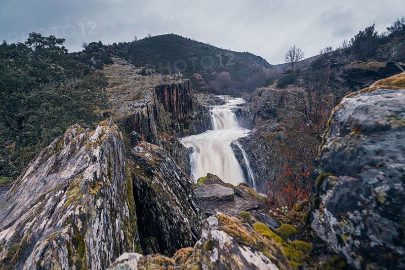 A majestic view of Cascada del Aljibe in Guadalajara, Spain, showcasing the powerful waterfall amidst rugged cliffs and lush vegetation, under moody skies, captured from an elevated perspective