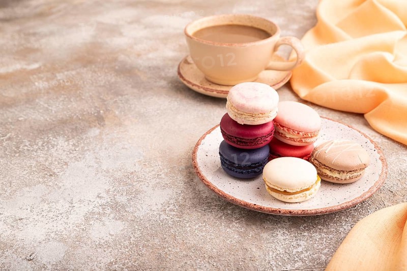 Macarons colourful cookies dessert, cup of coffee, on brown concrete background with orange linen textile, copy space, side view
