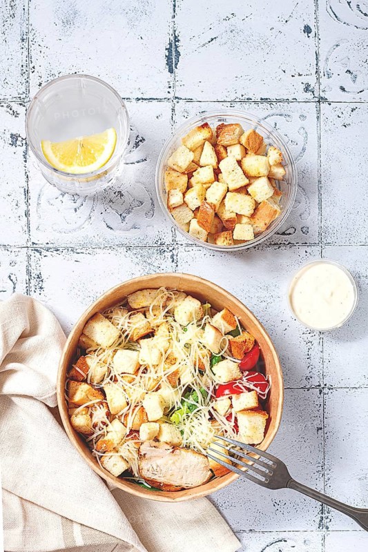 Caesar salad in a paper container, take it on the way, with sauce and croutons, on the table, snack, natural light, no people