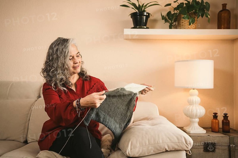 A woman with gray hair knits peacefully on a beige sofa, surrounded by plants and warm lighting. The cozy atmosphere highlights a sense of relaxation and creativity