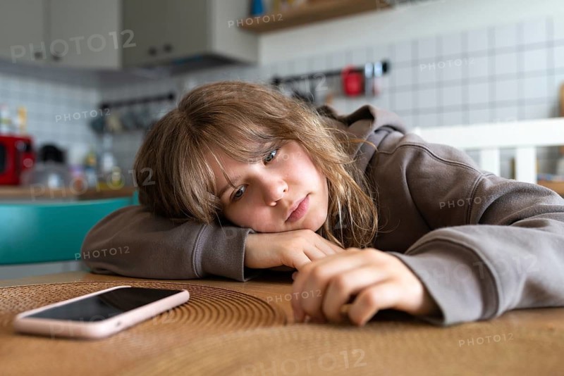 Teen girl rests her head on a table at home, deeply absorbed by her smartphone, highlighting her attachment to social media and technology. The phone is prominently placed beside her