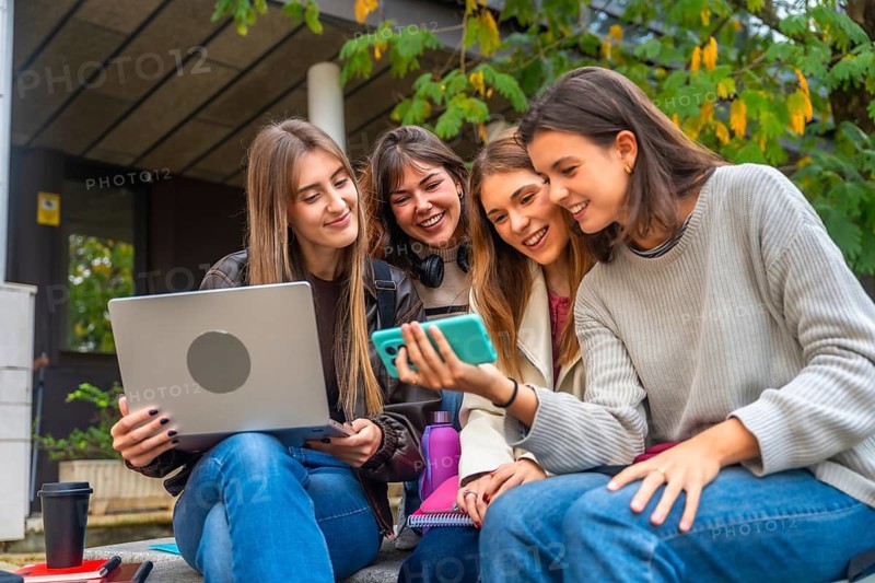 Group of female university students sitting outdoors, interacting with a smartphone and laptop, enjoying shared moments, and staying connected with digital tools
