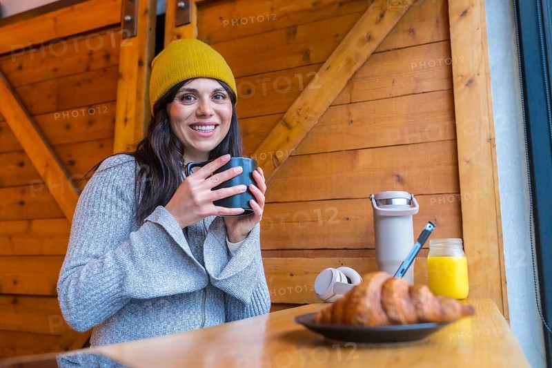 Woman holding a coffee mug, smiling at the camera, and enjoying a croissant, orange juice, and hot beverage in an urban cafe, reflecting a relaxed lifestyle on a cold city morning