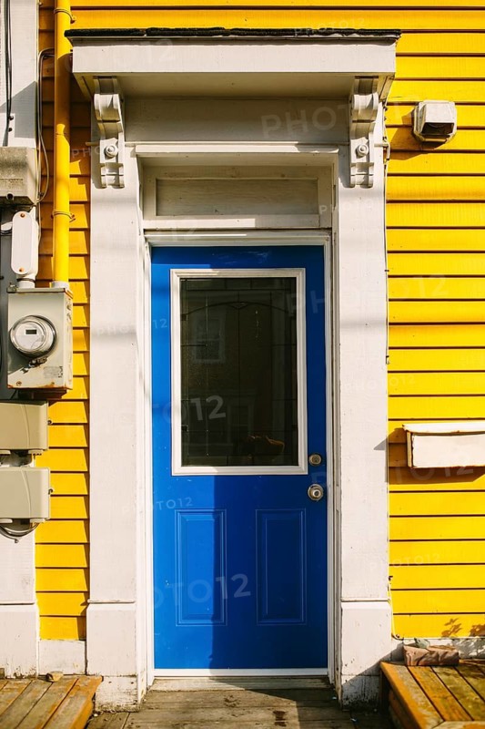 A striking bright blue door set against a vivid yellow wooden facade of a house in St. John's, Newfoundland & Labrador, Canada. The sunlit setting highlights the contrast between the colors and architectural details