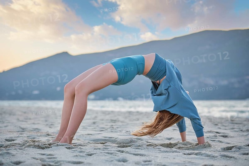 A woman practices Urdhva Dhanurasana on sandy beach at sunset with mountains in the background. She focuses on her yoga pose
