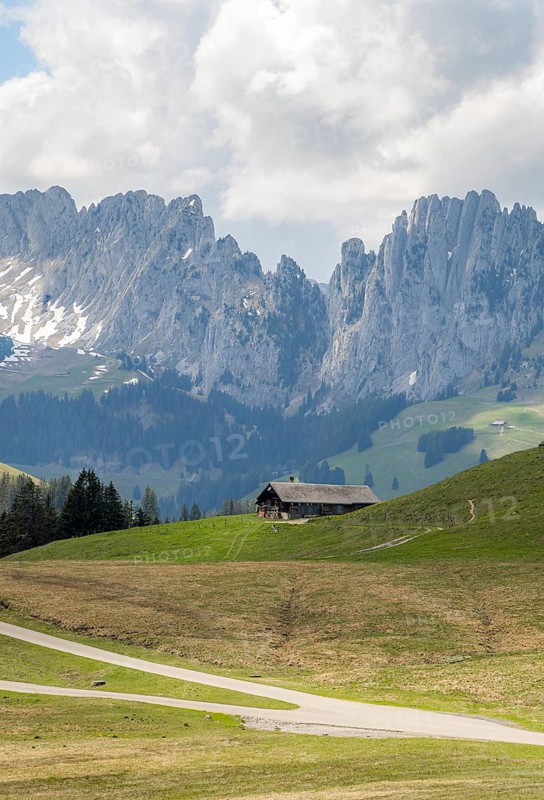 Spring view of the serene landscape in Jaun, located in the Gruyere district, Switzerland. A wooden house sits below the rugged peaks of the Gastlosen mountains, surrounded by lush green fields