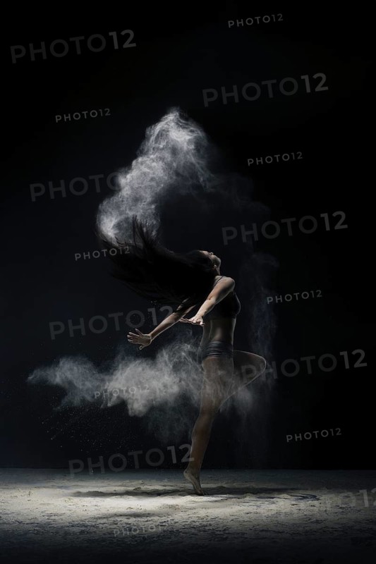 Brunette in black shorts and top jumping gracefully in a white dust cloud shot in dark room