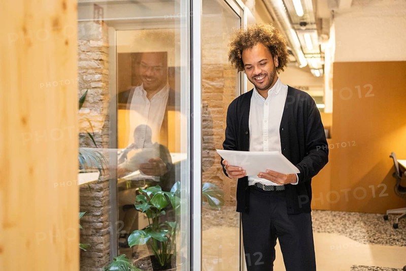 A man in a coworking space holds a document, smiling as he stands near a glass wall. The modern office interior features natural elements like plants and exposed brickwork