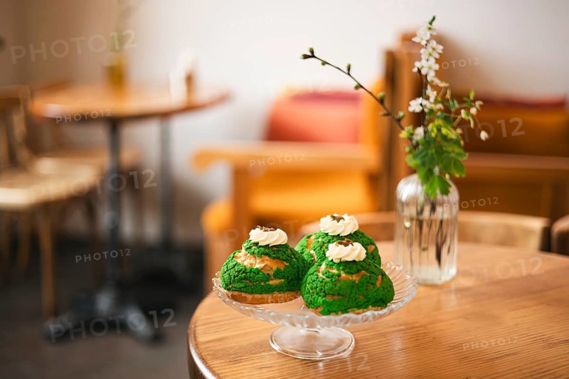 A glass plate with matcha choux pastries with whipped cream sits on a wooden table in a cozy cafe. In the background, blurred furniture and a flower vase add warmth