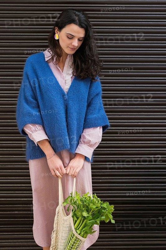 A woman wearing a blue cardigan and striped dress holds a mesh bag filled with fresh greens. Her colorful earrings add a vibrant touch, contrasting with the dark background