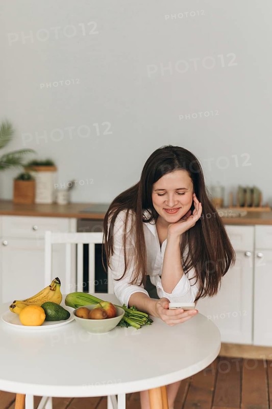A nutritionist is in a kitchen, smiling and using a smartphone. Fresh fruits and vegetables are on the table. The scene conveys healthy eating and wellness planning