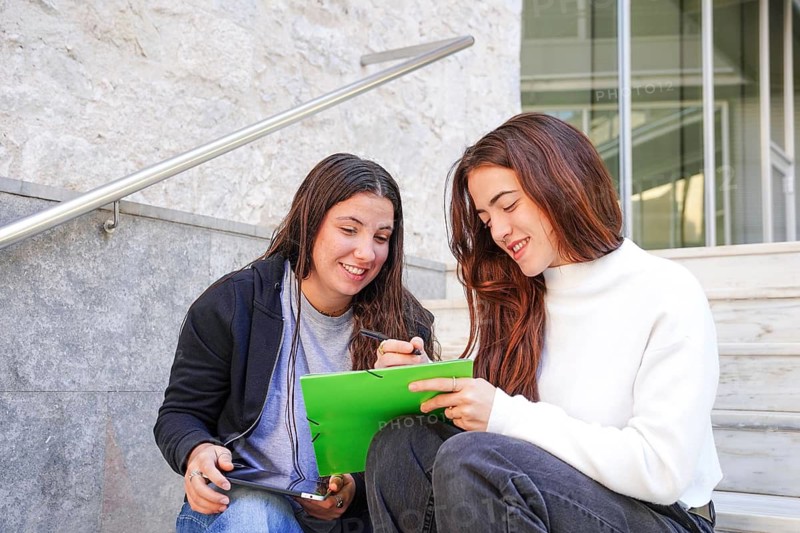 Two friends sitting on steps outdoors, one holding a bright green notebook and a pen, the other a tablet They are smiling and discussing, creating a positive, collaborative atmosphere
