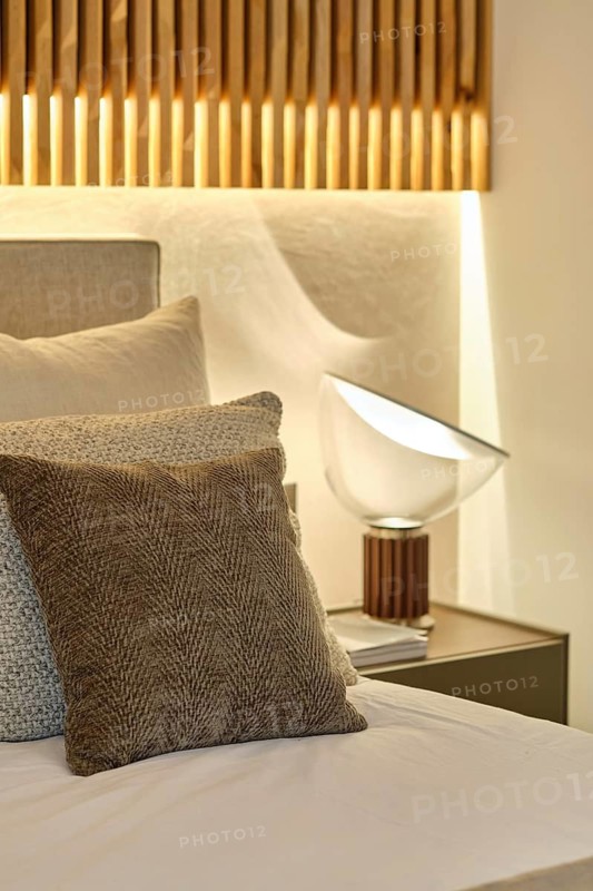 An elegant bedroom featuring a textured grey pillow, soft linen, a modern bedside lamp perched on a sleek table, and warm lighting emanating from a wooden slatted wall