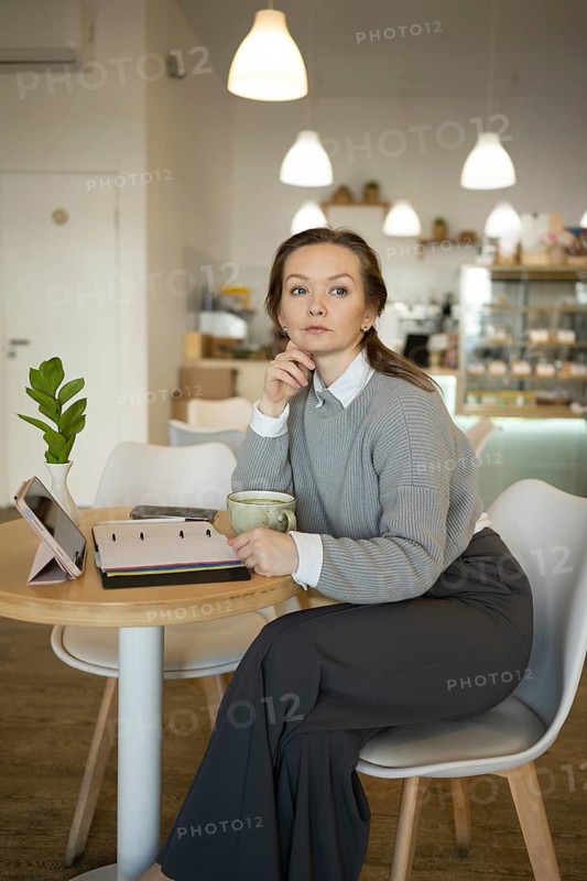 Woman sits at a cafe table with a coffee cup, tablet, and notebook. She appears thoughtful and organized, surrounded by a modern, cozy cafe ambiance