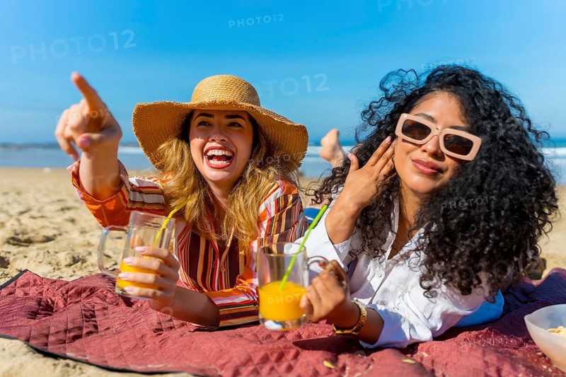 Two cheerful female friends relaxing on a picnic blanket by the ocean, holding refreshing drinks and pointing excitedly at something in the distance during their beach getaway