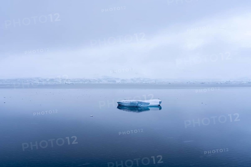 Small iceberg floating on mirror-like water surface, snow-covered coast in fog in the background, Wilhelmina Bay, Antarctic Peninsula, Antarctica