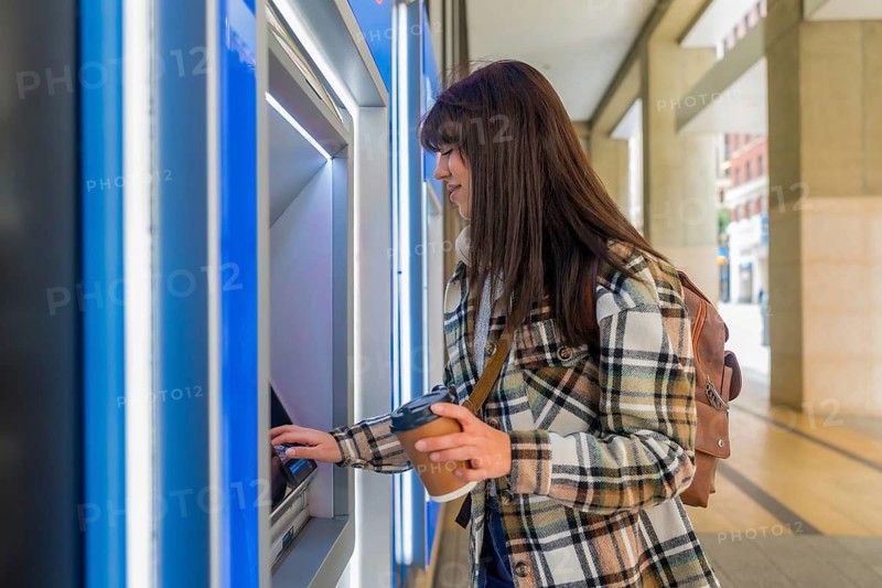 Woman standing at an automated teller machine, inserting a bank card into the slot while holding a reusable coffee cup, performing a banking transaction in an urban street