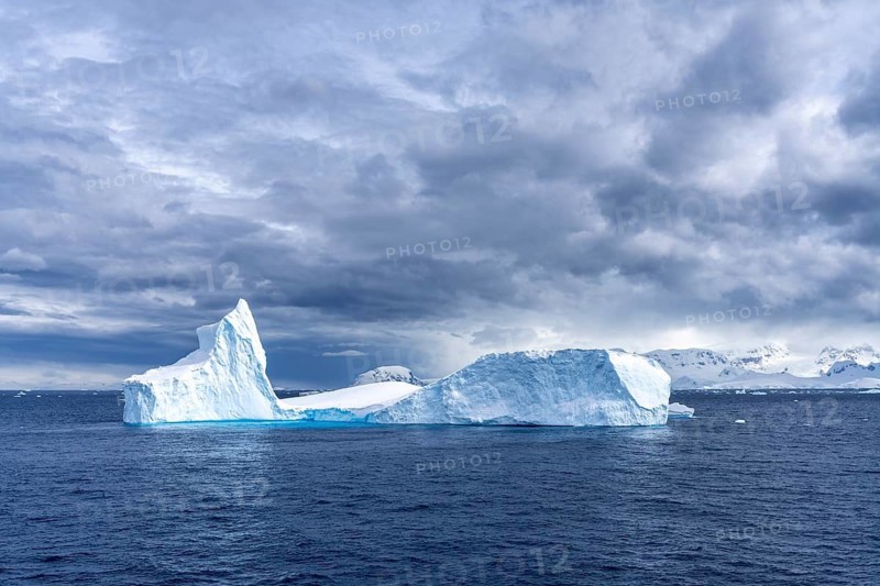 Iceberg with prominent tip and tabular section floating in the Southern Ocean, snow-covered mountain peaks in the background under overcast sky, Antarctic Peninsula, Antarctica