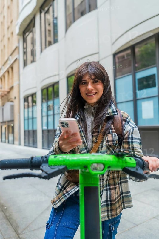 Woman standing on an electric scooter, navigating a city street and smiling at the camera while holding her smartphone, representing modern urban mobility, connectivity, and convenient travel