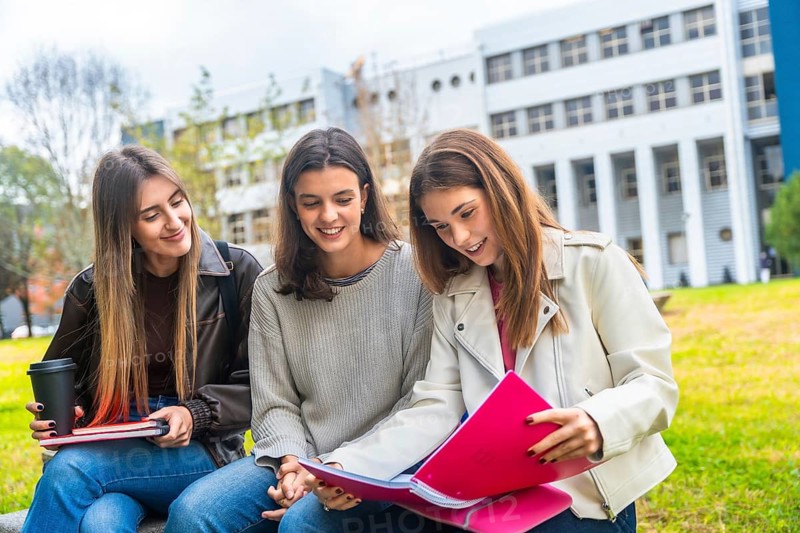 Three university students studying together outdoors on campus, sharing textbooks, coffee and notes while smiling, collaborating and preparing for exams in a relaxed, friendly setting