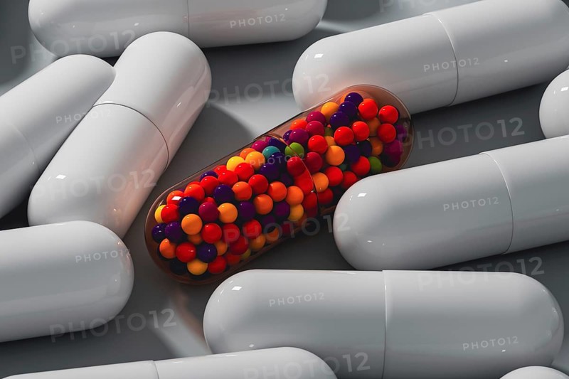 A vibrant capsule filled with colorful beads stands out among white medicine tablets on a gray surface, creating a striking contrast