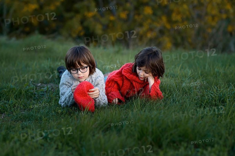 A young boy and girl, siblings, enjoy a leisure day in a grassy park surrounded by autumn colors. The boy, wearing glasses and a gray sweater, and the girl, in a bright red sweater, are laying on the grass