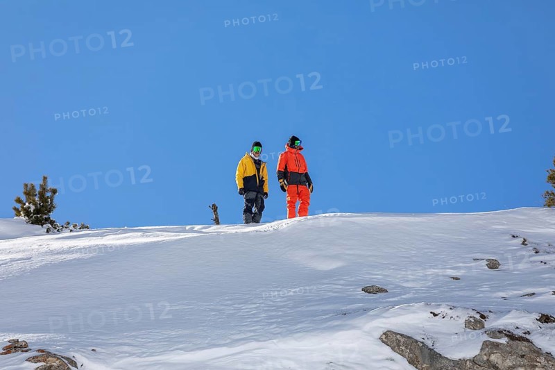 Two snowboarder friends in colorful winter gear stand on a snowy mountain slope, ready to descend. The clear blue sky and scattered pine trees enhance the alpine scene