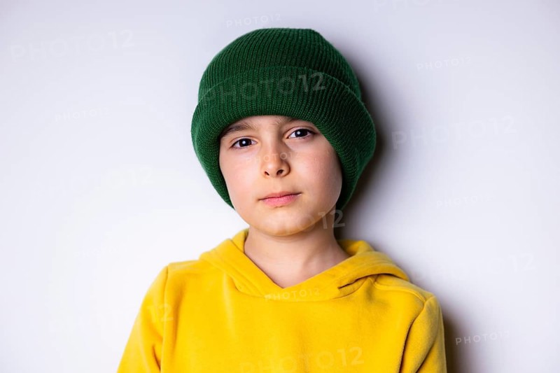 Portrait of a young child wearing a green knit beanie and a bright yellow hoodie, standing against a plain white background. The child looks directly at the camera with a calm expression