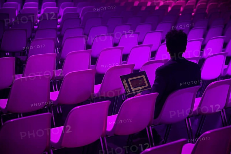 A indian man with a laptop sits amidst rows of empty chairs illuminated by purple lighting at a conference of AI in Amsterdam, suggesting focus and a modern digital workspace