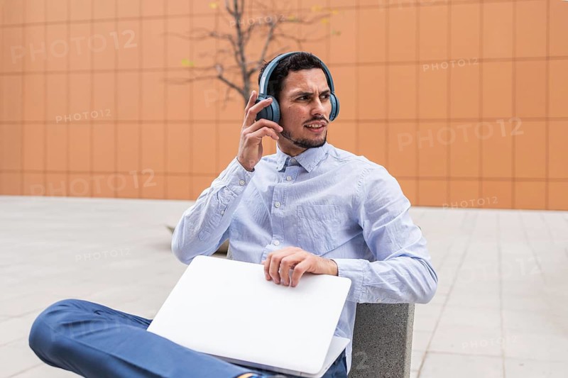 Man in business casual attire sits outside with a laptop and headphones, appearing focused while listening. The modern urban setting emphasizes productivity, remote work, and digital connectivity
