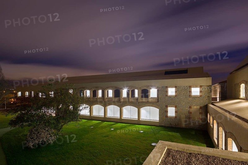 A modern hospital with stone facade and arched windows is lit from within against a twilight sky. The illuminated windows contrast with the tranquil, grassy surroundings