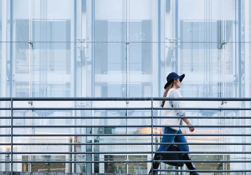 Side view of a woman in a stylish outfit and hat walking briskly against a backdrop of modern architecture. The image captures urban life and contemporary fashion