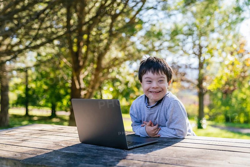A young boy with Down syndrome sits at a wooden table with a laptop, surrounded by lush greenery. His joyful expression reflects the excitement of learning in a natural outdoor setting