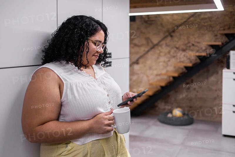A young woman enjoys a break in a modern coworking space, holding a coffee mug and using her smartphone, showcasing a blend of relaxation and technology in a casual work environment