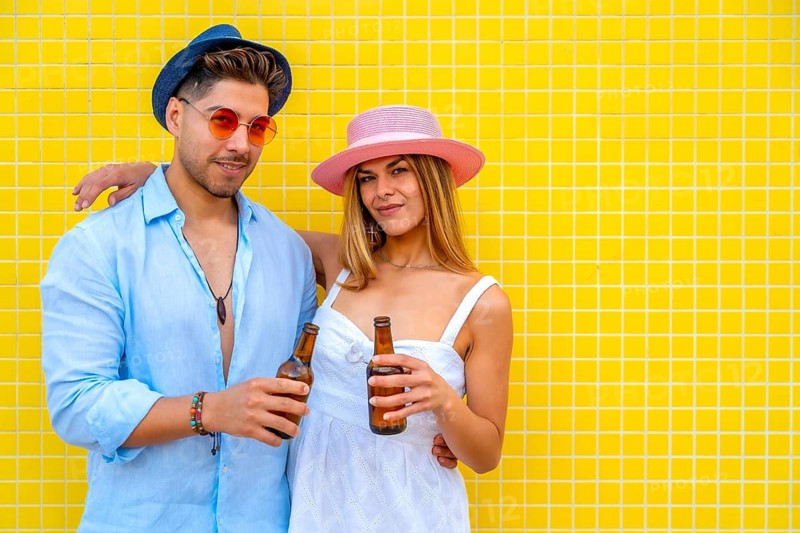 Young stylish couple holding beer bottles and smiling brightly in front of a vibrant yellow tiled wall, enjoying a carefree summer day together