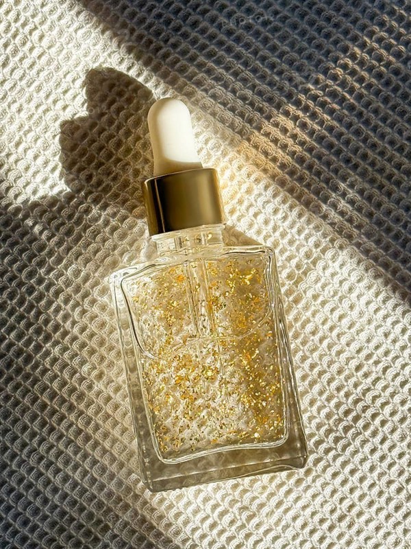 A transparent glass dropper cosmetic bottle filled with a luxurious serum containing gold flakes, set on textured fabric in the bathroom. Sunlight casts warm shadows, enhancing the elegant scene